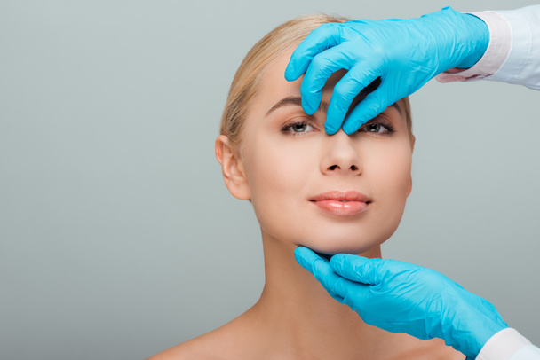 cropped view of beautician in latex gloves touching nose of happy naked woman isolated on grey  - Fotoğraf, Görsel