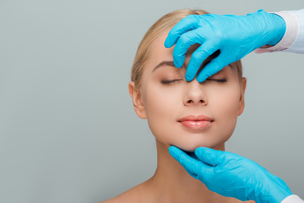 cropped view of beautician in latex gloves touching nose of naked woman isolated on grey  - Photo, Image