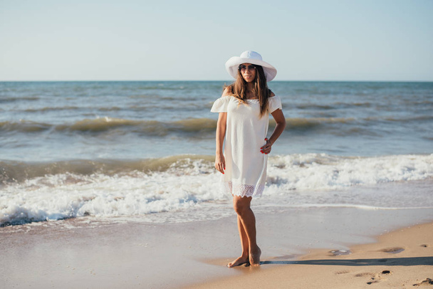 όμορφη ευτυχισμένη νεαρή γυναίκα σε λευκό φόρεμα, καπέλο και γυαλιά ηλίου με τα πόδια στην αμμώδη παραλία - Φωτογραφία, εικόνα