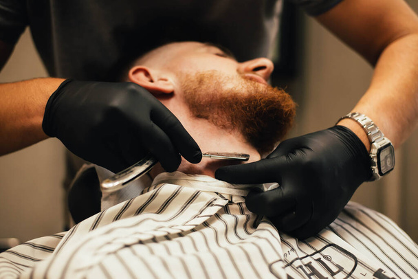 cropped shot of barber shaving beard to man in barbershop - Fotó, kép