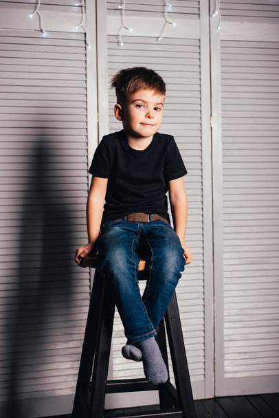 adorable little boy in black t-shirt and jeans sitting on stool and looking at camera - Fotoğraf, Görsel