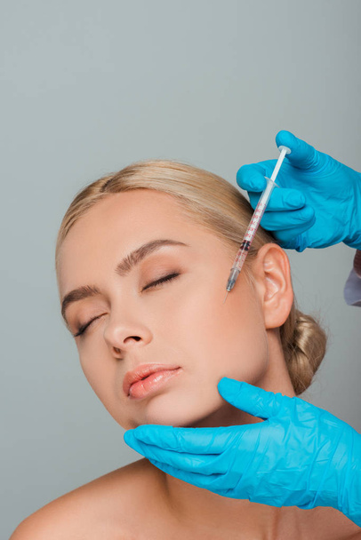 Ausgeschnittene Ansicht einer Kosmetikerin in Latex-Handschuhen mit Spritze in der Nähe der Wange einer jungen Frau isoliert auf grau  - Foto, Bild