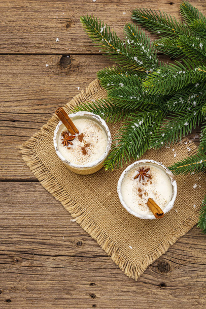 Eggnog cocktail in two glasses arranged with christmas decoration on old wooden table. Evergreen fer tree branch, artificial snow, sackcloth napkin - Photo, Image