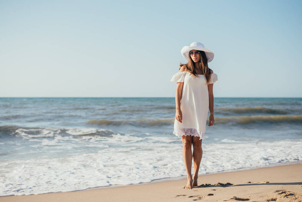 beautiful happy young woman in white dress, hat and sunglasses walking on sandy beach - 写真・画像