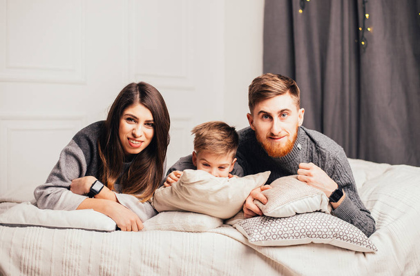 1人の子供を持つ幸せな若い家族がベッドに横になりカメラで笑っています  - 写真・画像