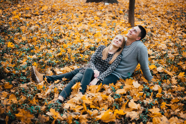 happy young couple sitting together on yellow leaves in autumn forest - Photo, Image