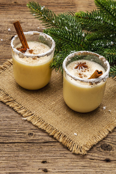 Eggnog cocktail in two glasses arranged with christmas decoration on old wooden table. Evergreen fer tree branch, artificial snow, sackcloth napkin - Φωτογραφία, εικόνα