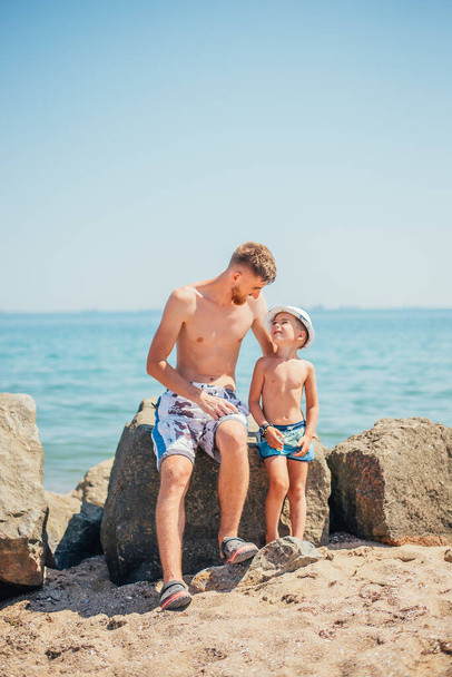 happy father and son in swimwear sitting together on rock on beach - Фото, изображение