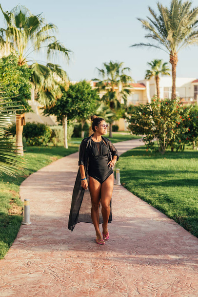 beautiful young woman walking on walkway between palm trees at tropical resort - Foto, Imagen