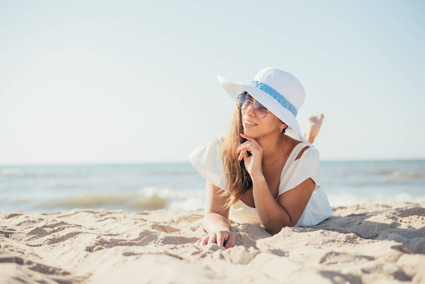 attractive young woman in hat and sunglasses lying on sandy beach at sunny summer day - Photo, image