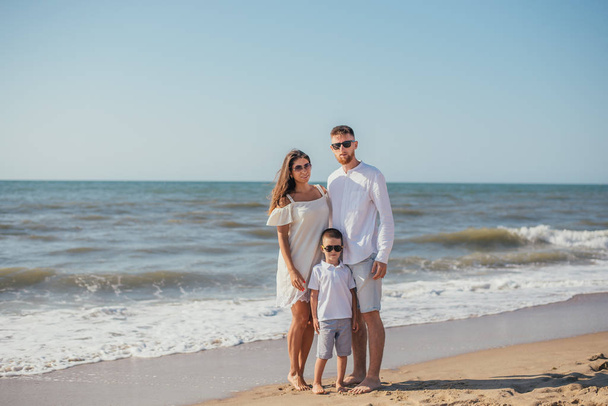 feliz familia joven con un niño de pie juntos y sonriendo a la cámara en la playa de arena en verano
 - Foto, Imagen