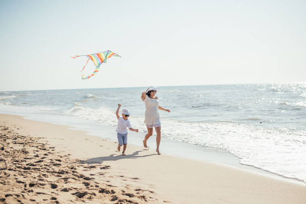 happy mother and son playing with colorful kite on sandy beach at summertime - Фото, изображение