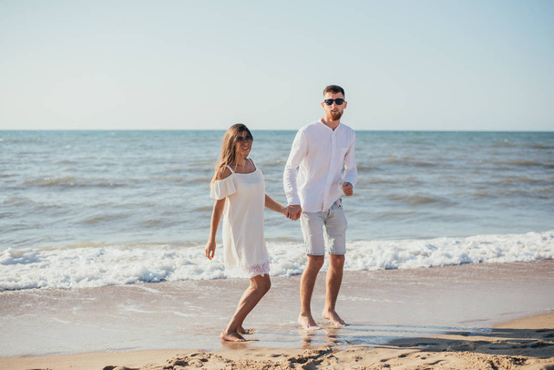 happy young couple holding hands and standing together on sandy beach at summertime - Фото, изображение