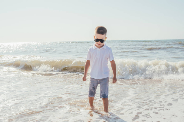 cute happy boy in shorts and sunglasses walking on sandy beach  - Fotografie, Obrázek