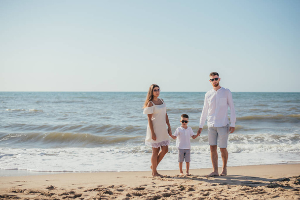счастливая молодая семья с одним ребенком, стоящая вместе и держащаяся за руки на песчаном пляже в летнее время
 - Фото, изображение