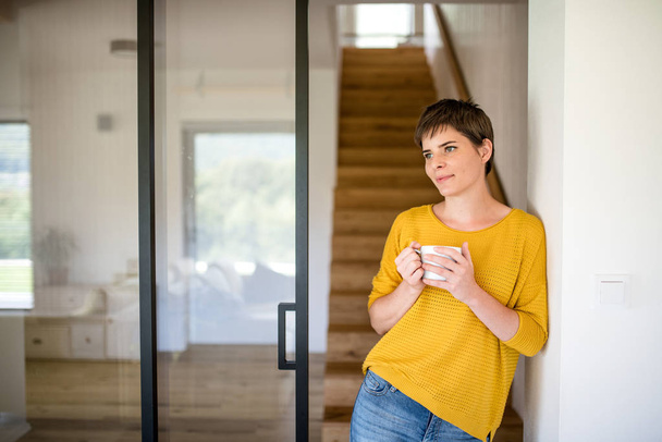 Portrait of young woman with coffee standing indoors at home, leaning on wall. - Foto, Imagen