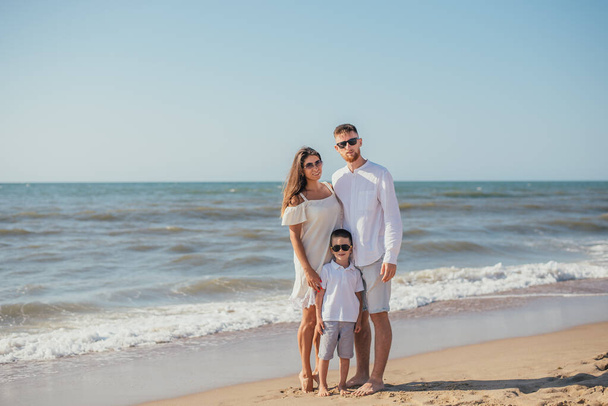 szczęśliwa młoda rodzina z jednym dzieckiem stojącym razem na piaszczystej plaży latem - Zdjęcie, obraz