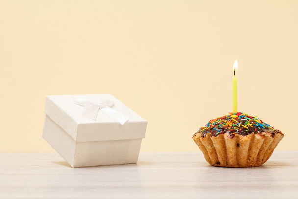 Tasty birthday muffin with burning festive candle and gift box on beige background. - Photo, Image