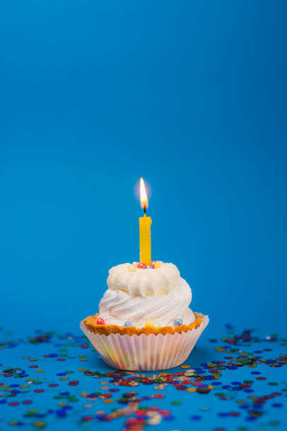 Крупный план вкусного праздничного кекса с горящей свечой и красочными конфетти на голубом фоне
 - Фото, изображение
