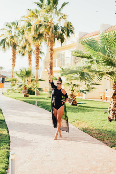 attractive young woman walking on walkway between palm trees at tropical resort - Fotó, kép