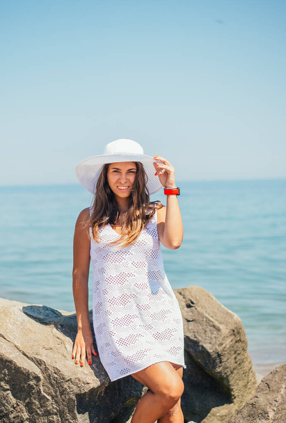 beautiful happy young woman in white dress and hat posing on rocks at sea - Фото, изображение