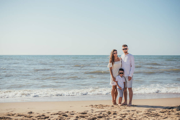 счастливая молодая семья с одним ребенком, стоящая вместе и улыбающаяся фотоаппарату на песчаном пляже летом
 - Фото, изображение