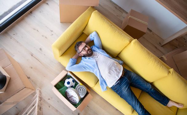 Vista superior del hombre maduro con cajas moviéndose en casa nueva, relajándose en el sofá
. - Foto, Imagen