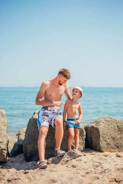 happy father and son in swimwear sitting together on rock on beach  - Foto, Imagem
