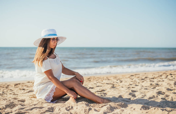 attractive young woman in hat and sunglasses sitting on sandy beach at summertime  - Valokuva, kuva