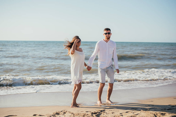 happy young couple holding hands and standing together on sandy beach at summertime - 写真・画像