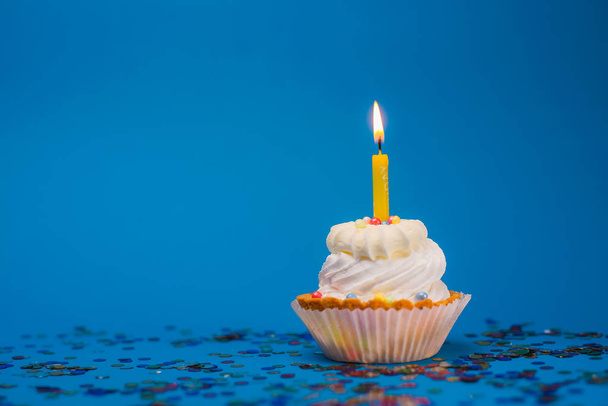 close-up uitzicht van heerlijke feestelijke cupcake met brandende kaars en kleurrijke confetti op blauwe achtergrond - Foto, afbeelding