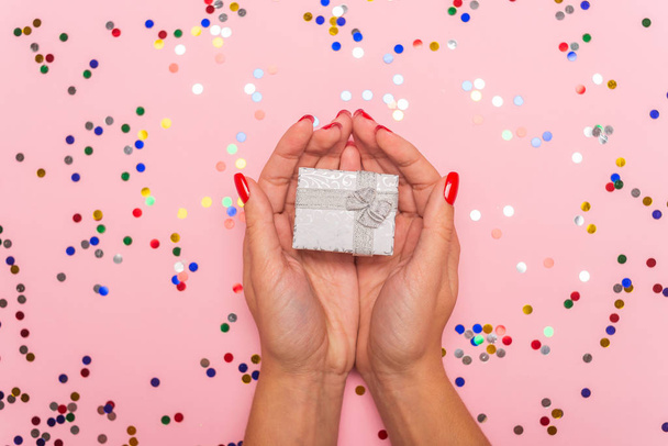 close-up partial view of woman holding gift box on pink background with colorful confetti - Foto, imagen