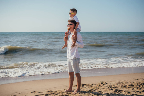 счастливый молодой отец, несущий на шее очаровательного маленького сына на песчаном пляже летом
 - Фото, изображение