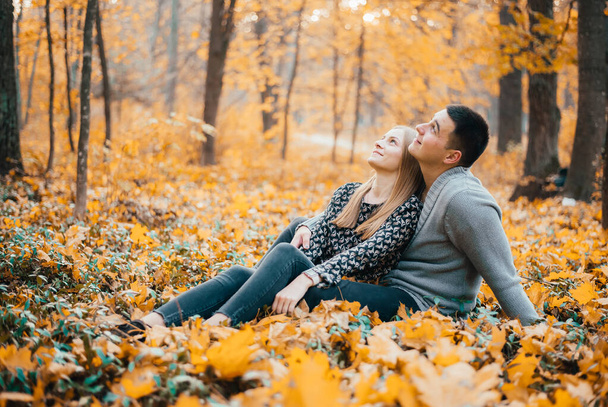 happy young couple sitting together on yellow leaves in autumn forest - 写真・画像