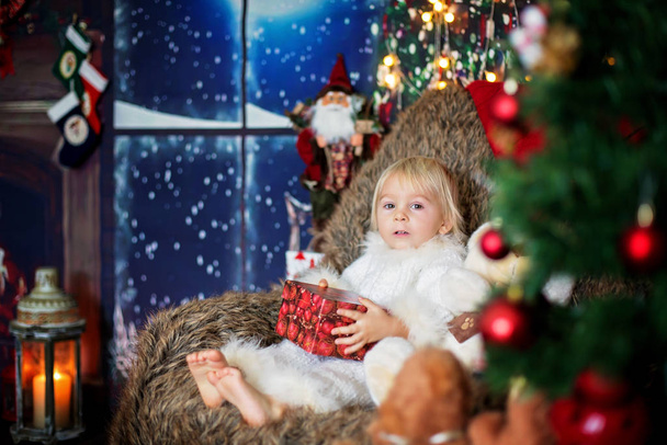 Blond toddler child, boy, opening present at home, sitting in co - 写真・画像
