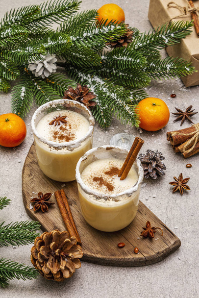 Eggnog with cinnamon and nutmeg for Christmas and winter holidays. Homemade beverage in glasses with spicy rim. Tangerines, candles, gift. Stone concrete background - Photo, image