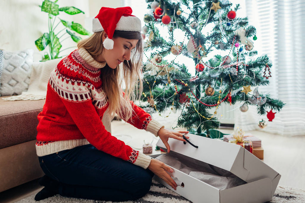Weihnachtsmorgen und Neujahrsgeschenke. Frau mit Weihnachtsmannhut öffnet Geschenkbox am Weihnachtsbaum zu Hause - Foto, Bild