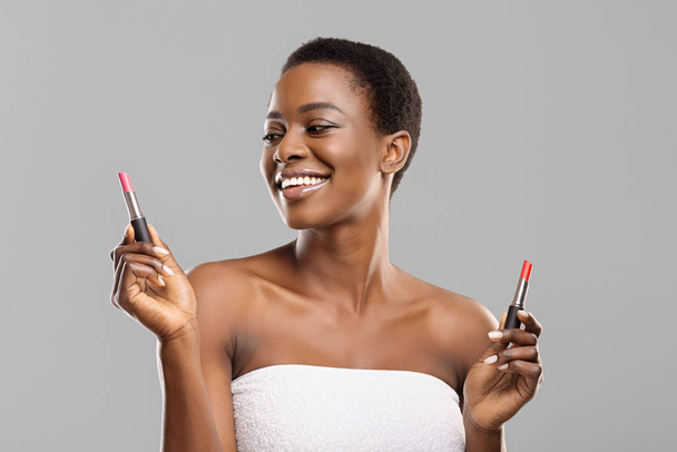 Radosna afro kobieta trzyma dwie szminki, wybierając właściwy kolor - Zdjęcie, obraz
