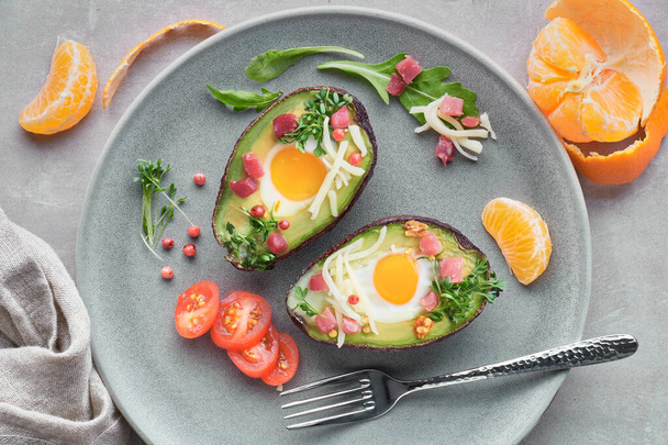 Keto diet dish: baked Avocado boats with ham cubes, quail eggs and cheese served with salad on a gray table with a drink and mandarines - Foto, Bild