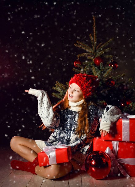 Fiatal lány ül ünneplő téli karácsonyfa díszített vörös fejjel megható hópelyhek hó alatt - Fotó, kép
