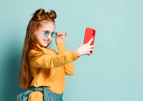 Ένα όμορφο κοκκινομάλλικο κορίτσι βγάζει μια selfie με ένα νέο κινητό τηλέφωνο σε μια κόκκινη θήκη. Σε μπλε γυαλιά και ένα κίτρινο πουλόβερ - Φωτογραφία, εικόνα