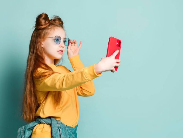 美しい赤い髪の少女は、赤いケースに新しい携帯電話で自撮りを取ります.青い眼鏡と黄色のセーターで - 写真・画像
