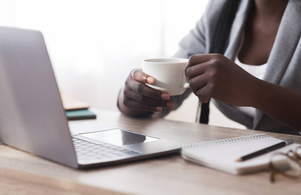 Unrecognizable black businesswoman drinking coffee and using laptop in office - Foto, imagen