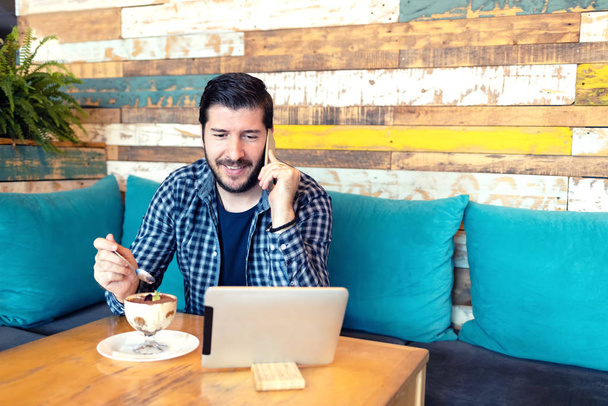 erfolgreicher Mann mittleren Alters, der im Café telefoniert, während er sein digitales Tablet benutzt und im trendigen Café Dessert genießt - Fernbedienung - Foto, Bild