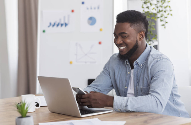 Jovem empresário sorrindo usando smartphone e trabalhando no laptop no escritório
 - Foto, Imagem