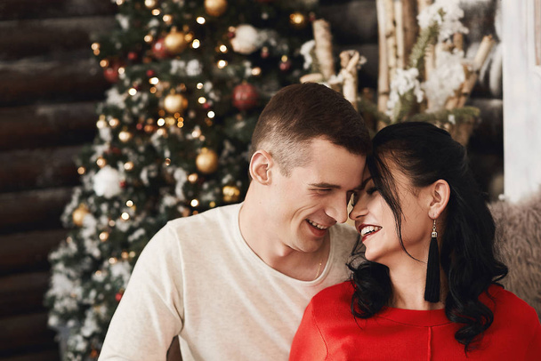 Close up portrait of a young beautiful couple of lovers, happy brunette girl in red dress and smiling handsome man posing in an interior with festive Christmas lights on the background. - Zdjęcie, obraz
