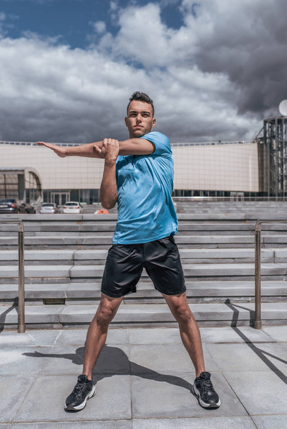 Male athlete, warms up muscles, workout training, arm and shoulder muscles, summer in city. Fitness motivation youth lifestyle. Sportswear, shorts and T-shirt in sneakers. Cloud stair background. - Photo, Image