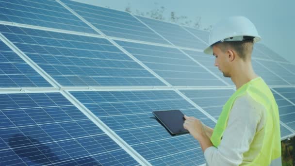 Side view: Worker in overalls and a helmet works with a tablet on the background of ground-based solar panels. Alternative energy - Felvétel, videó