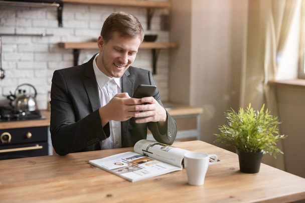 young smiling businessman reading something funny in his smartphone in the kitchen while drinking morning coffee before going to office. multitasking, morning - Foto, immagini