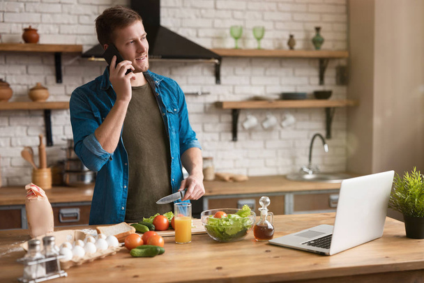 Mutfakta kahvaltı hazırlarken telefonda konuşan gülümseyen genç adam pazar sabahı laptopunda tarifini kontrol ediyor. - Fotoğraf, Görsel